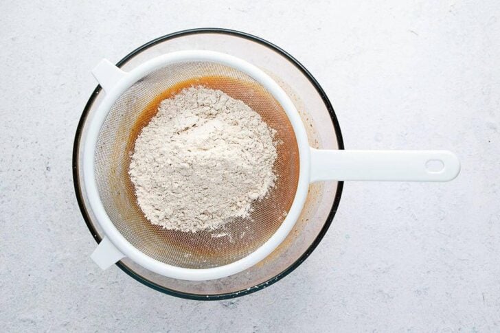 A metal mesh sieve with flour sits atop a glass mixing bowl on a light surface, ready for sifting.
