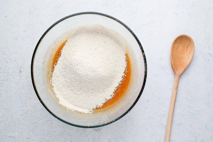 A glass mixing bowl with flour on top of wet ingredients sits on a light surface next to a wooden spoon.