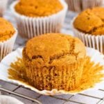 A golden-brown muffin with a cracked top sits on an opened white paper liner, surrounded by other muffins in liners, all resting on a cooling rack.