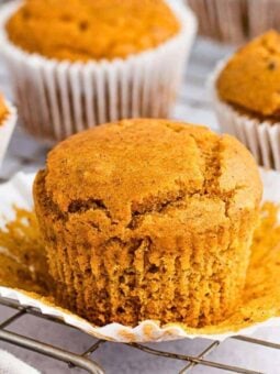 A golden-brown muffin with a cracked top sits on an opened white paper liner, surrounded by other muffins in liners, all resting on a cooling rack.