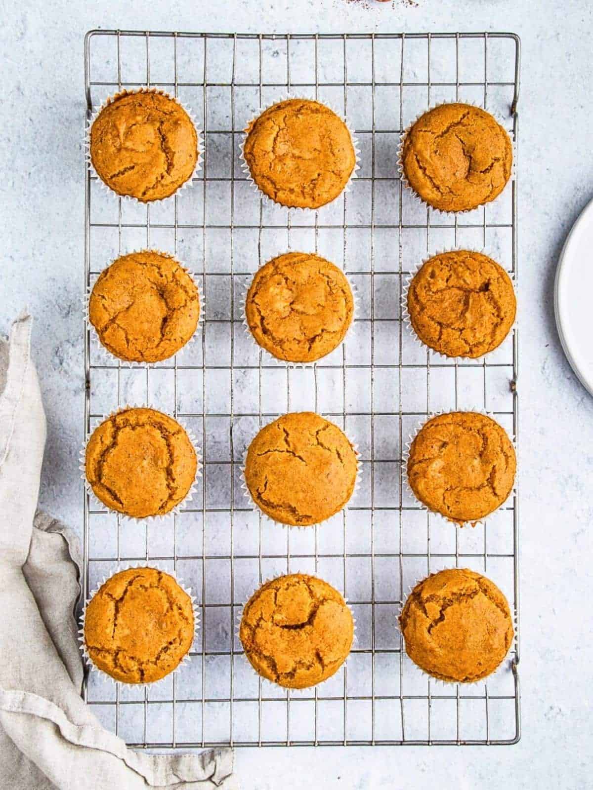Golden brown muffins arranged in three rows on a wire cooling rack, set on a light textured surface. A beige cloth is partially visible on the left and a white plate appears on the right edge of the image.