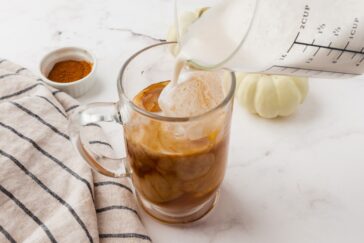 A glass mug filled with iced coffee is being topped with creamy milk poured from a measuring cup. A bowl of cinnamon and a striped cloth napkin are beside the mug on a white surface.