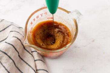 A green-handled whisk blends a brown liquid in a Pyrex measuring cup beside a striped cloth on a white surface.