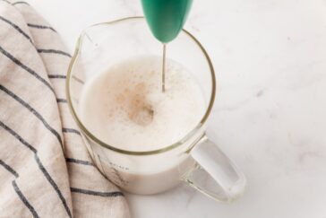 A handheld milk frother is frothing milk in a glass measuring cup on a white surface beside a striped kitchen towel.