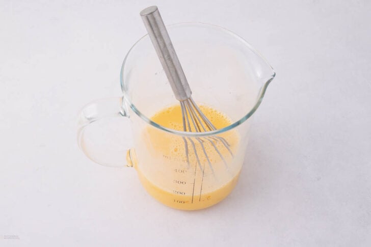 A glass measuring cup filled with a yellow liquid mixture, likely beaten eggs, sits on a white surface. A metal whisk rests inside the cup.