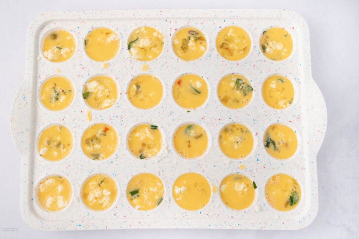 A speckled white muffin tin filled with uncooked egg mixture, vegetables, and cheese, ready to be baked, sits on a light surface.