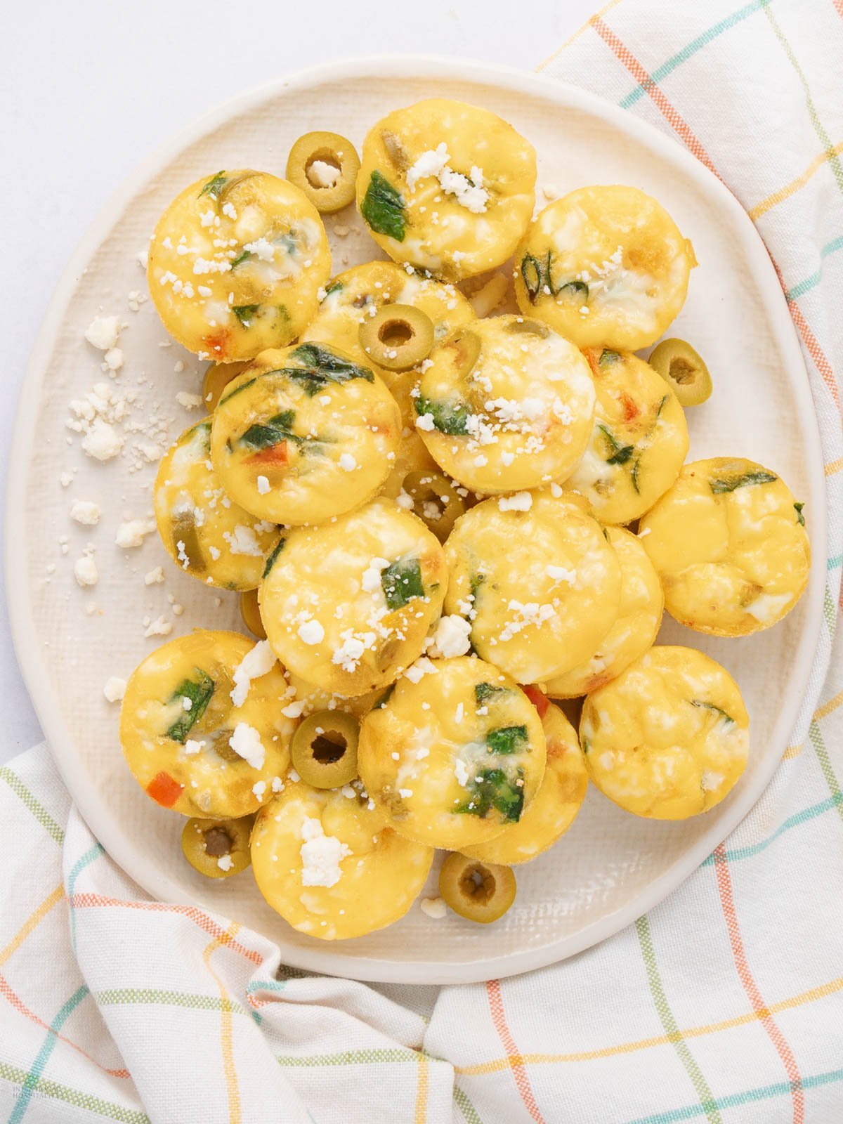 A plate of mini egg muffins with vegetables and green olives, topped with crumbled cheese, on a white dish with a plaid napkin underneath.