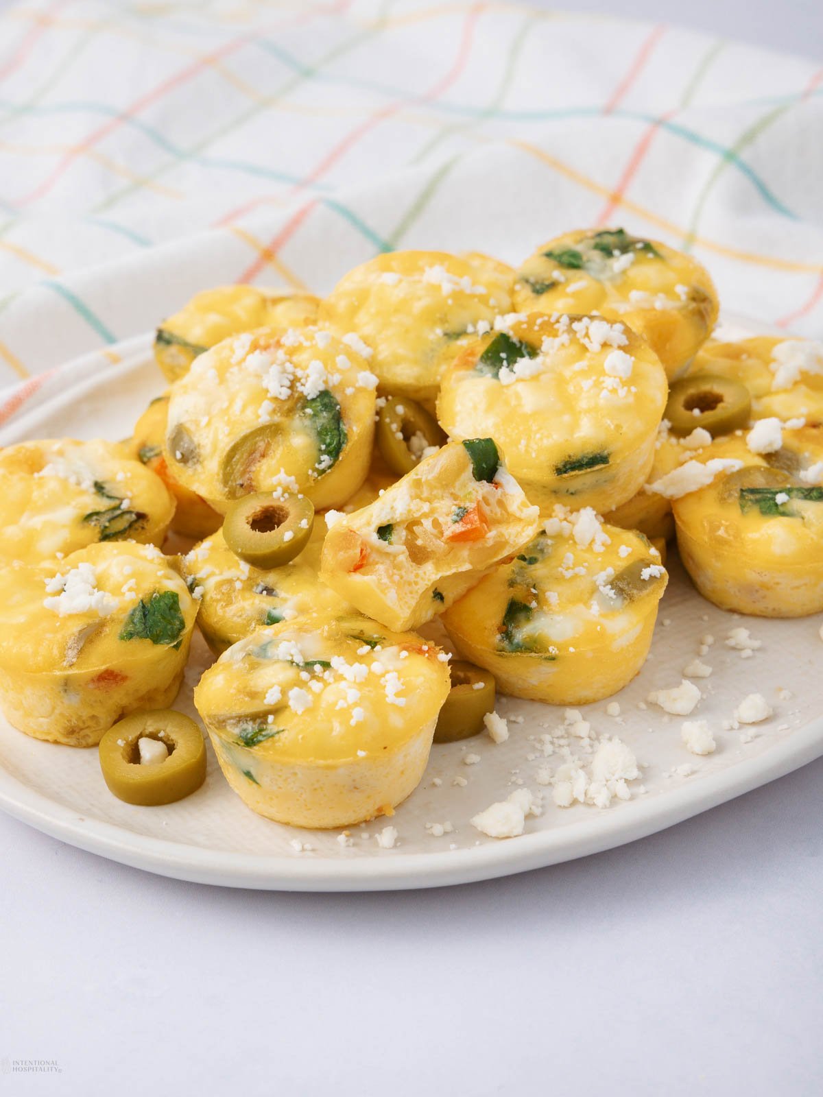 A plate of mini egg muffins with sliced green olives, spinach, and crumbled cheese on top, arranged on a white plate with a colorful checkered cloth in the background.