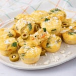 A plate of mini egg muffins with spinach and cheese, topped with crumbled feta and sliced green olives, sits on a white plate. A colorful, plaid-patterned cloth is in the background.