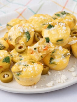 A plate of mini egg muffins with spinach and cheese, topped with crumbled feta and sliced green olives, sits on a white plate. A colorful, plaid-patterned cloth is in the background.