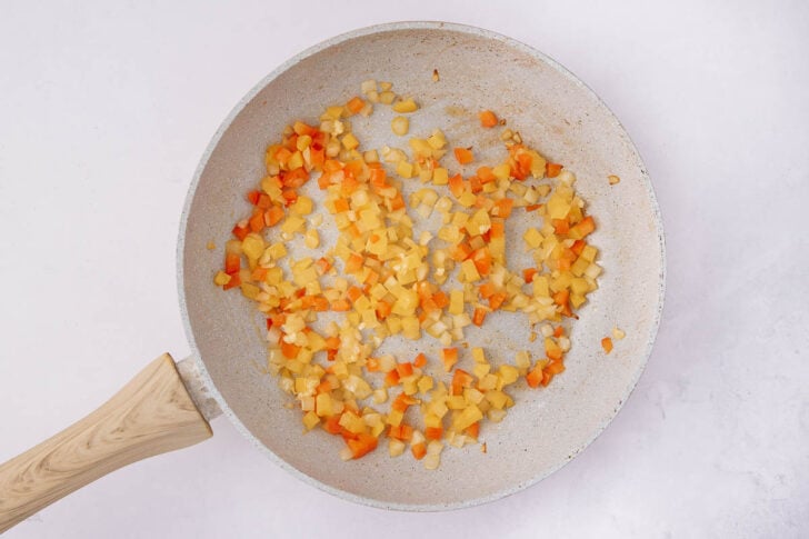 A frying pan with diced yellow potatoes and orange bell peppers being sautéed, viewed from above on a light surface. The vegetables are partially cooked and spread across the pan.