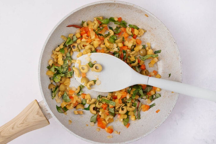 A white frying pan with a wooden handle contains chopped vegetables, including green olives, red peppers, onions, and leafy greens. A white spatula rests in the pan, mixing the colorful ingredients.