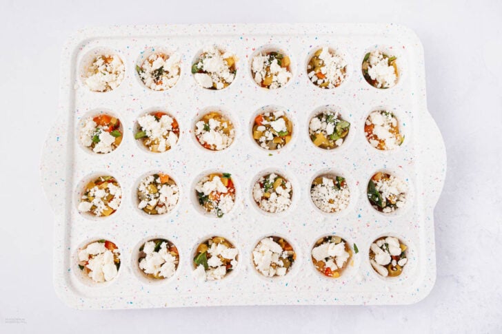 A white 24-cup muffin tray filled with uncooked mini egg bites topped with crumbled feta cheese, mixed vegetables, and herbs, set on a light background.