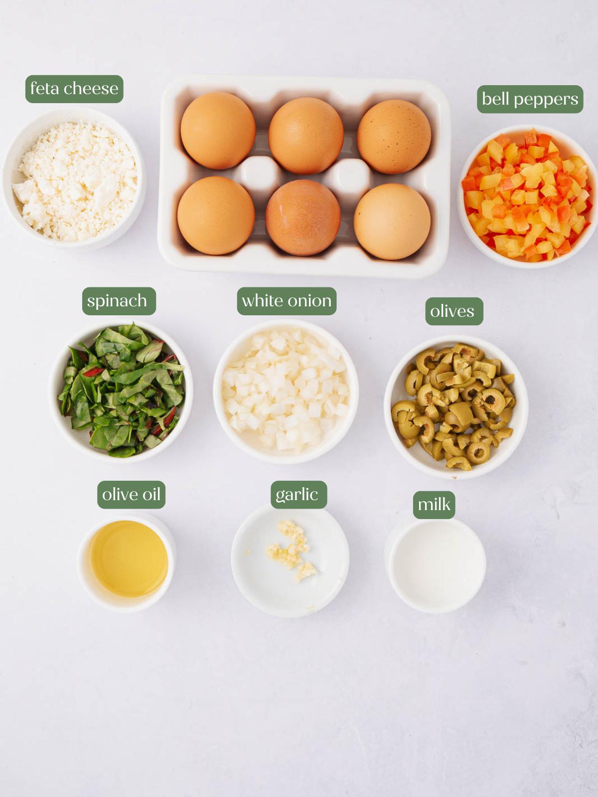 A flat lay of ingredients in bowls labeled: feta cheese, eggs, diced bell peppers, chopped spinach, diced white onion, sliced olives, olive oil, minced garlic, and milk on a white surface.