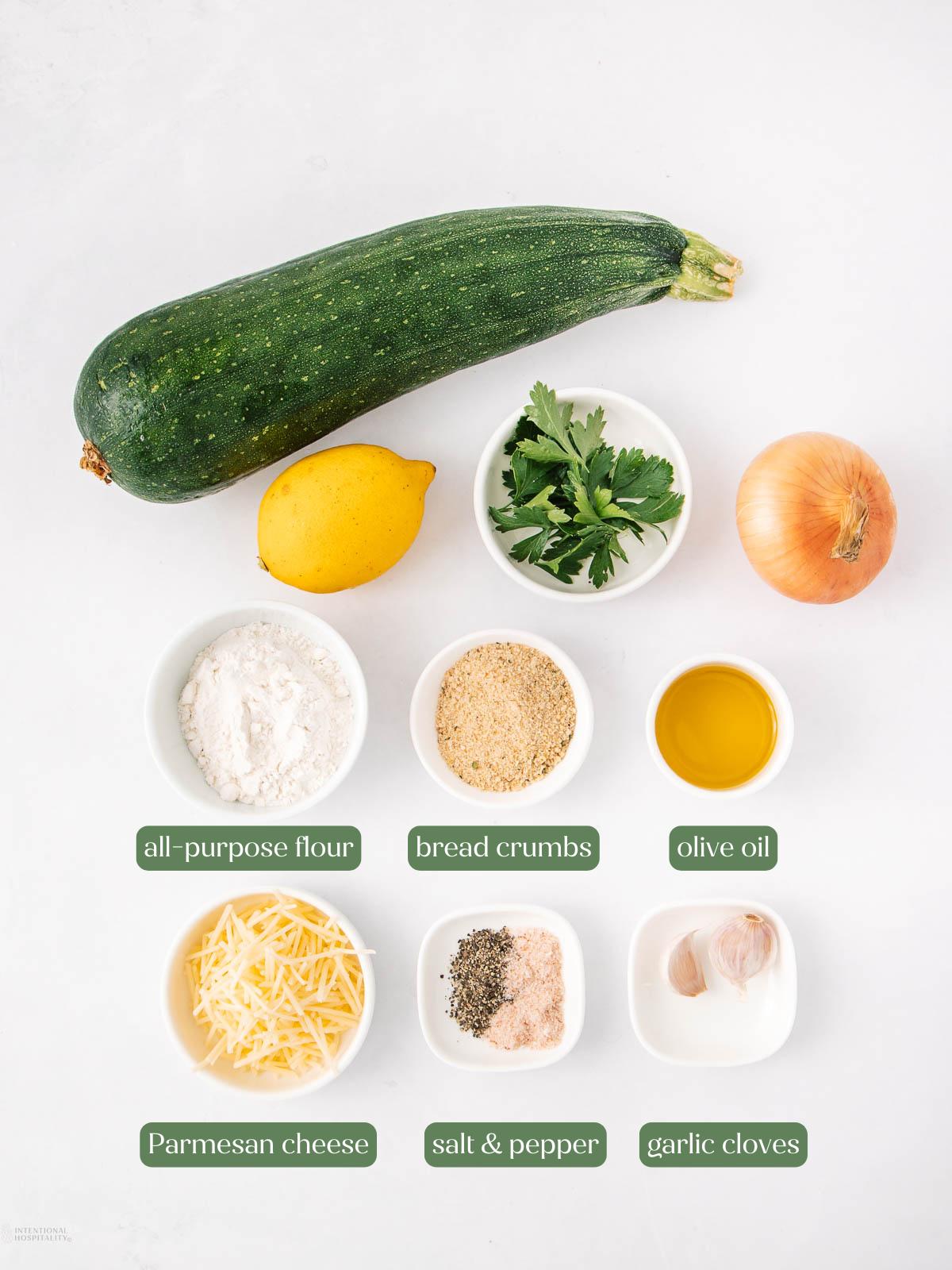 A zucchini, lemon, parsley, onion, olive oil, bread crumbs, all-purpose flour, Parmesan cheese, salt and pepper, and two garlic cloves are arranged on a white surface in small bowls.