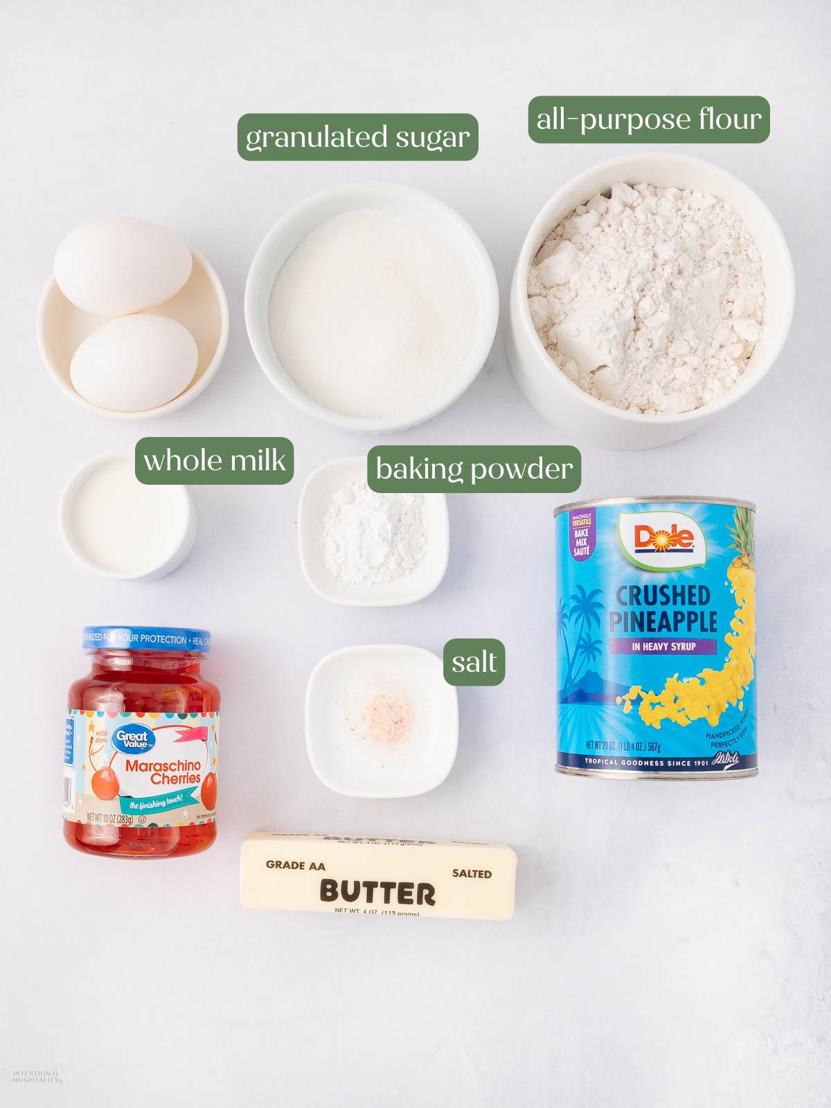 Labeled baking ingredients on a white surface: two eggs, granulated sugar, all-purpose flour, whole milk, baking powder, salt, a can of crushed pineapple, a jar of maraschino cherries, and a stick of salted butter.