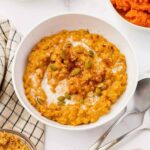 A bowl of pumpkin oatmeal topped with pumpkin seeds, a swirl of cream, and a sprinkle of cinnamon sits on a white surface next to a spoon, a striped cloth, and small bowls of ingredients.