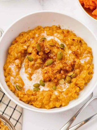 A bowl of pumpkin oatmeal topped with pumpkin seeds, a swirl of cream, and a sprinkle of cinnamon sits on a white surface next to a spoon, a striped cloth, and small bowls of ingredients.