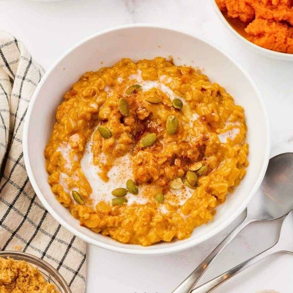 A bowl of pumpkin oatmeal topped with pumpkin seeds, a swirl of cream, and a sprinkle of cinnamon sits on a white surface next to a spoon, a striped cloth, and small bowls of ingredients.