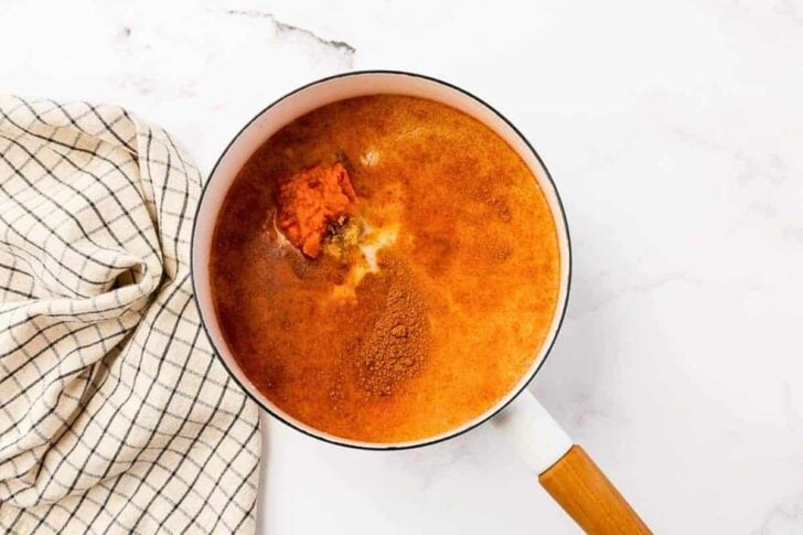 A white saucepan with a wooden handle contains a mixture of spices and tomato paste in liquid, next to a white and black checkered kitchen towel on a white surface.