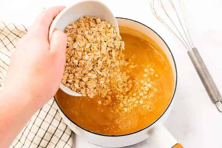 A hand pours rolled oats from a white bowl into a pot of simmering liquid. A whisk and a checkered kitchen towel are nearby on a white countertop.