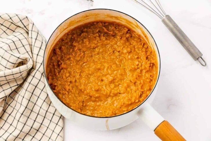 A white saucepan filled with thick, orange-brown cooked lentils or dal sits on a white surface next to a checkered kitchen towel and a metal whisk.