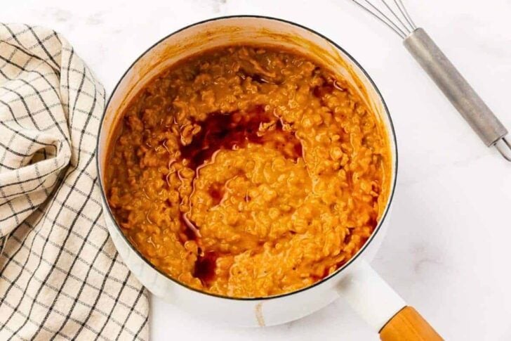 A white saucepan filled with creamy, orange-brown dal topped with a swirl of dark oil or sauce, next to a checkered cloth and a whisk on a white surface.