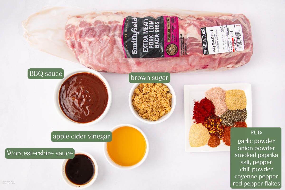 A rack of raw pork ribs with labeled ingredients: BBQ sauce, brown sugar, apple cider vinegar, Worcestershire sauce, and a bowl of spices for rub including garlic, onion, paprika, chili, cayenne, salt, pepper, and red pepper flakes.