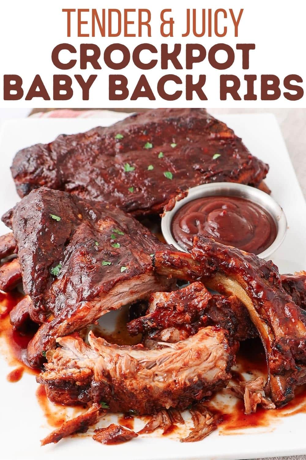 A plate of saucy, tender baby back ribs with chopped herbs on top, served with a small cup of barbecue sauce. The ribs are partially pulled apart to reveal juicy, cooked meat. Text above reads: "Tender & Juicy Crockpot Baby Back Ribs.