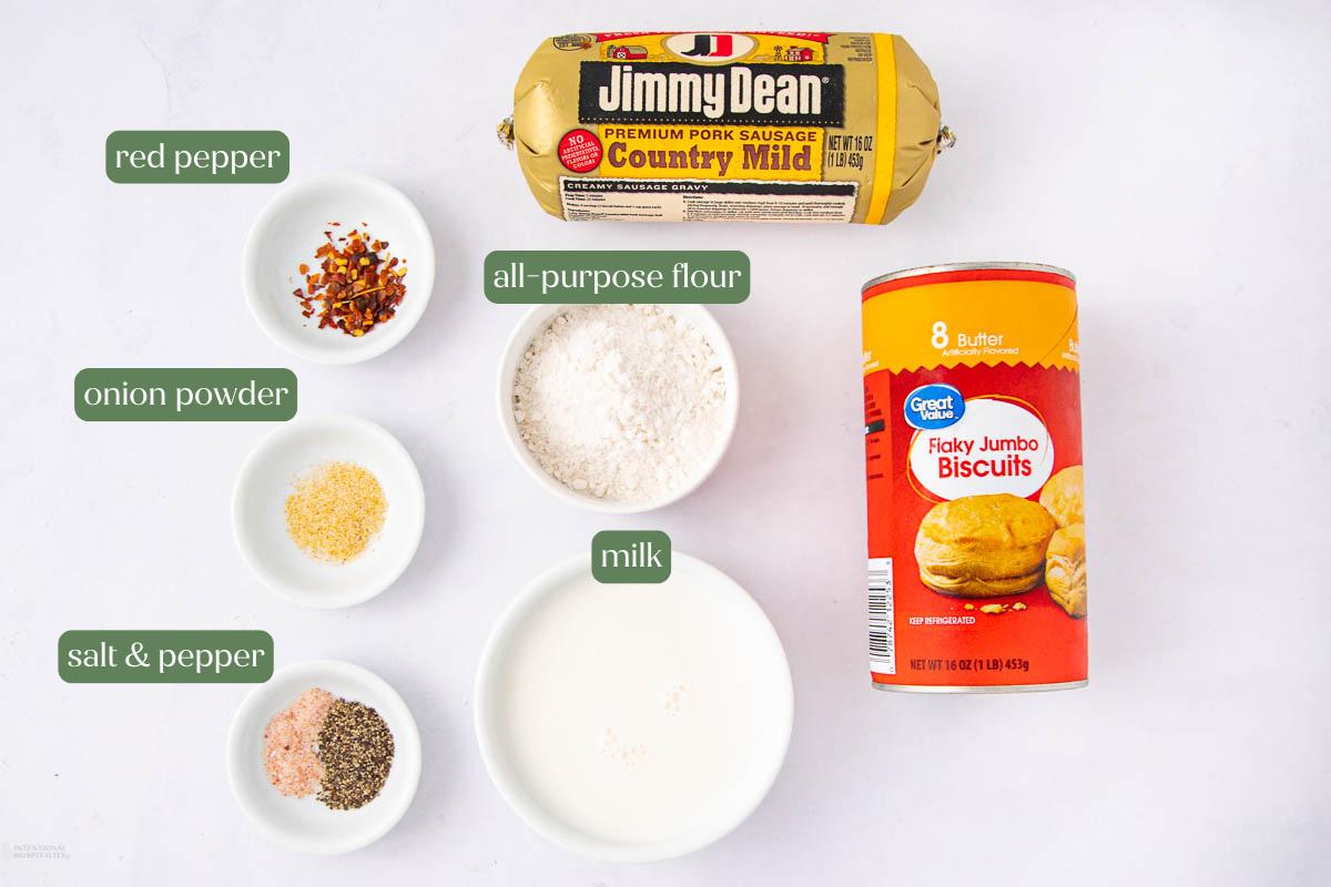 Ingredients for a recipe on a white background: a roll of Jimmy Dean pork sausage, a can of Great Value jumbo biscuits, small bowls of red pepper flakes, all-purpose flour, onion powder, milk, salt, and black pepper.