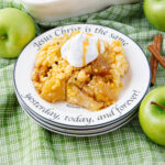 A plate of apple crisp topped with whipped cream sits on a green checkered cloth, surrounded by green apples and cinnamon sticks. The plate is inscribed with the Bible verse, "Jesus Christ is the same yesterday, today, and forever!.
