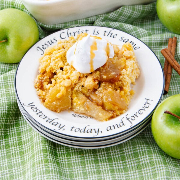 A plate of apple crisp topped with whipped cream sits on a green checkered cloth, surrounded by green apples and cinnamon sticks. The plate is inscribed with the Bible verse, "Jesus Christ is the same yesterday, today, and forever!.