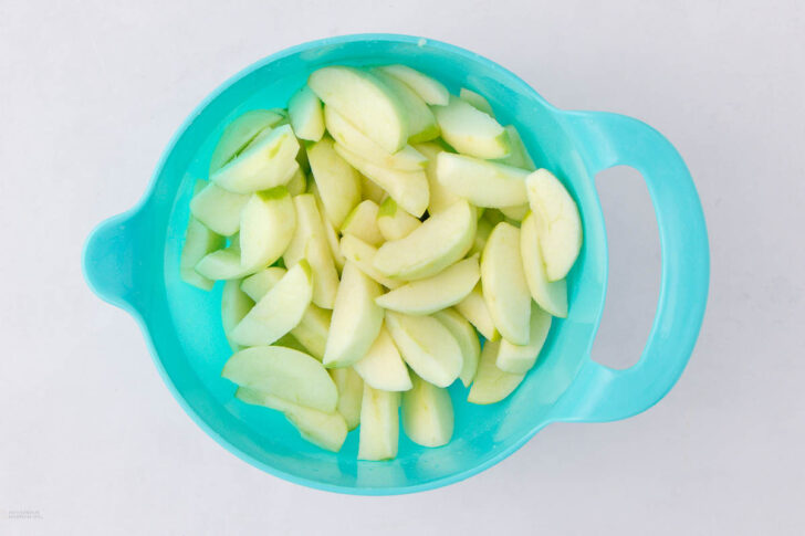 A blue bowl filled with peeled and sliced green apples sits on a white surface.