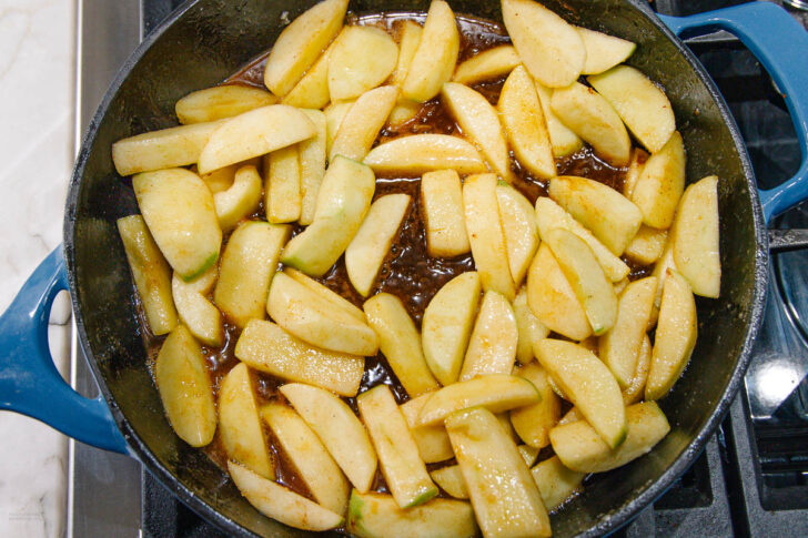 Sliced apples cooking in a blue cast iron skillet with a brown sugar and cinnamon sauce on a stovetop.