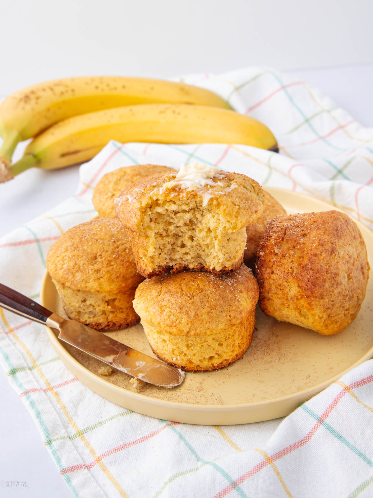 A plate of six golden-brown banana muffins, one split open with butter melting on top. A knife rests on the plate, and ripe bananas sit on a checkered cloth in the background.