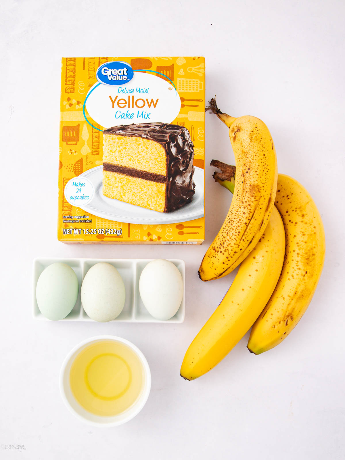 A box of yellow cake mix, three bananas, three eggs in a dish, and a small bowl of oil are arranged on a white background.