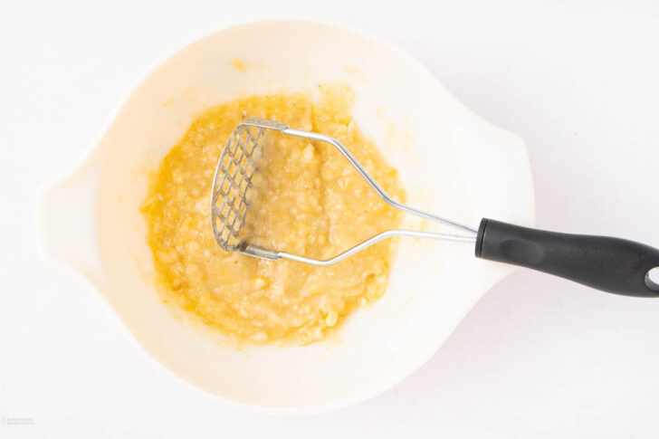 A white mixing bowl containing mashed bananas, with a metal potato masher featuring a black handle resting inside the bowl.