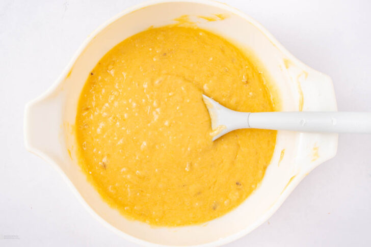 A white mixing bowl filled with banana bread batter, with a white spatula resting in the batter on a white surface.