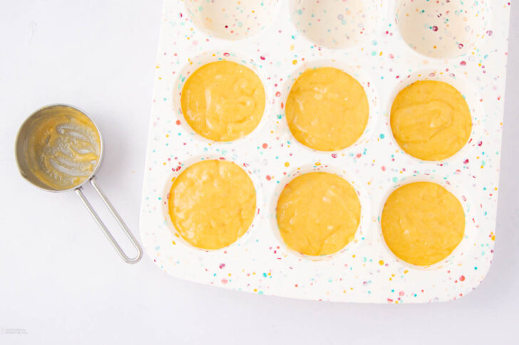 A white, speckled muffin tin filled with yellow batter sits on a white surface. Next to it is a metal measuring cup with some leftover batter inside.