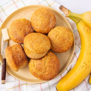 A plate of six golden brown muffins arranged in a circle, next to a butter knife with a pat of butter and two ripe bananas on a checkered cloth.