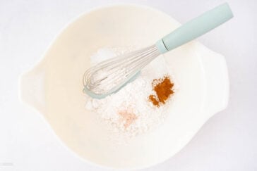 A mixing bowl with flour, ground cinnamon, and a pinch of pink salt, with a whisk resting on top of the ingredients.