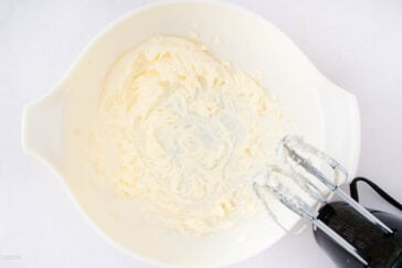 A white mixing bowl containing whipped cream cheese being blended with an electric hand mixer on a white surface.