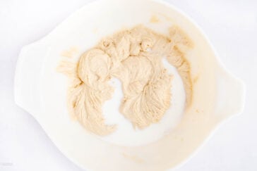 A white mixing bowl containing partially mixed light brown batter and some unmixed milk, creating a swirling pattern. The bowl is set against a white background.