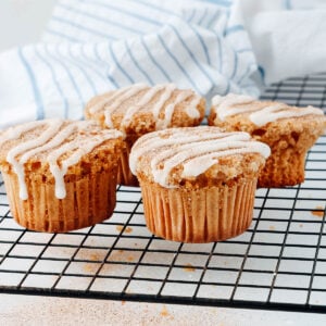 Four cinnamon sugar muffins with white icing drizzle are displayed on a cooling rack, with a striped kitchen towel in the background.