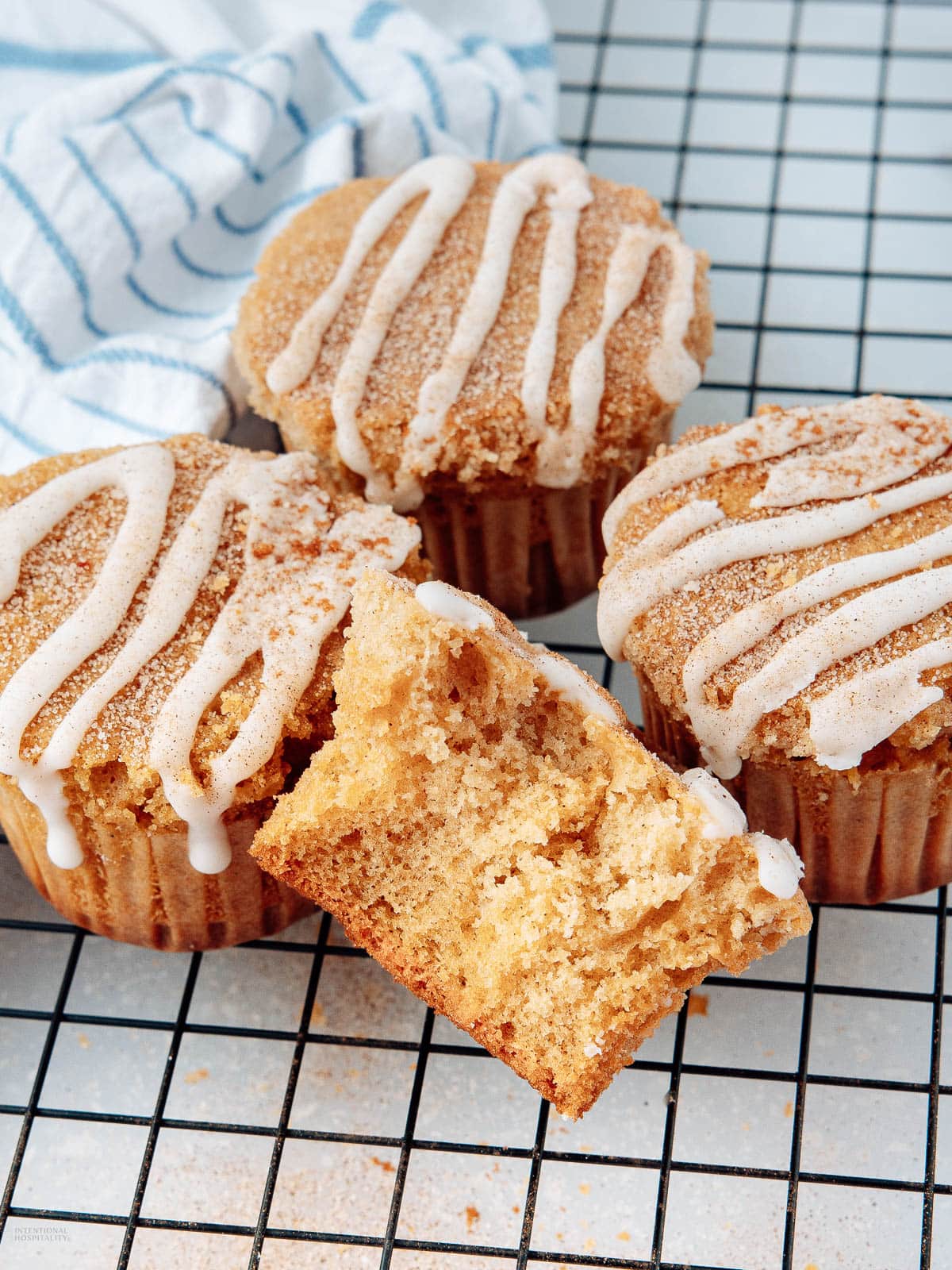 Three cinnamon muffins with white icing drizzle sit on a cooling rack. One muffin is broken in half, showing its moist, fluffy interior. A blue and white striped cloth is in the background.