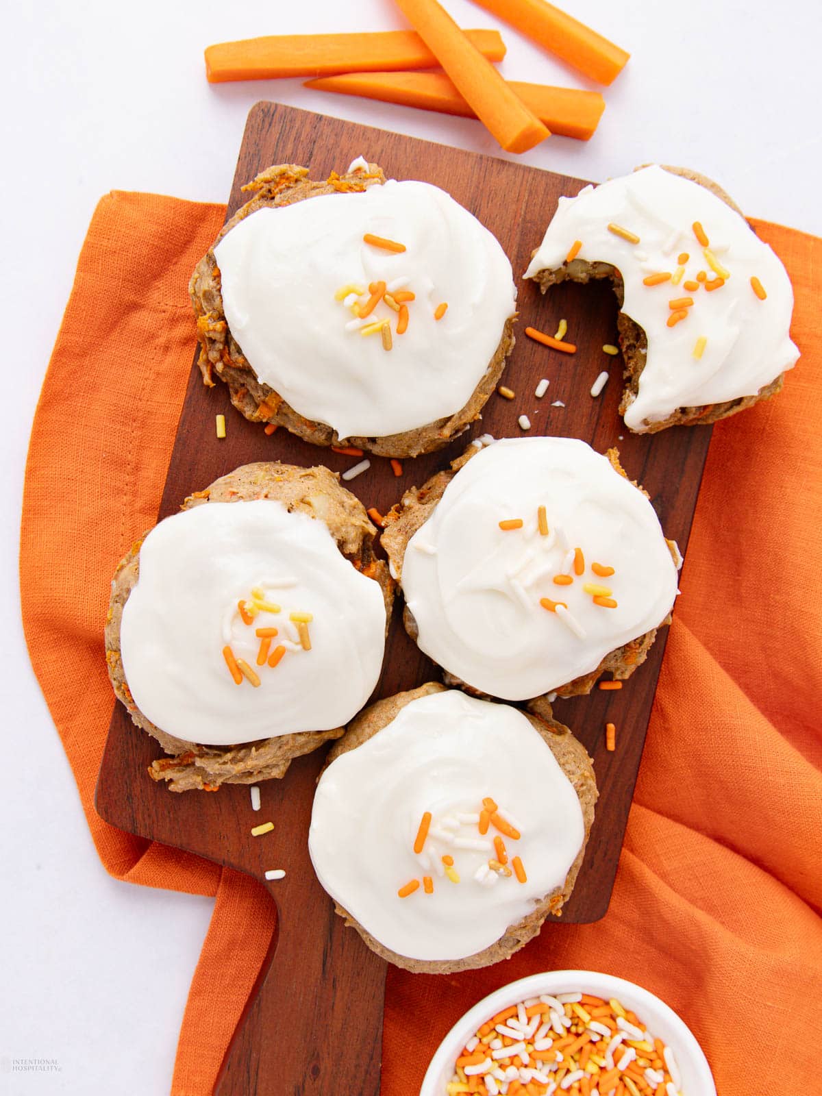 Five frosted cookies with orange and white sprinkles sit on a wooden board atop an orange cloth. One cookie has a bite taken out. Carrot sticks and a bowl of sprinkles are nearby.