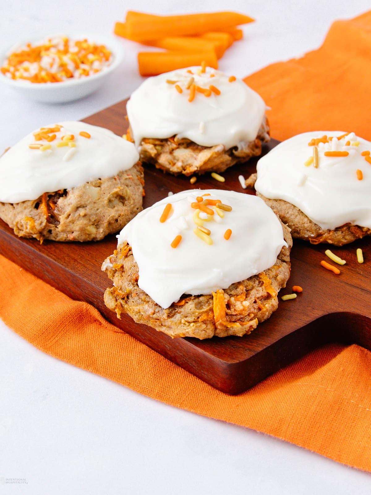 Four carrot cake cookies topped with white icing and orange sprinkles are arranged on a wooden board. An orange napkin and a small bowl of sprinkles and carrot sticks are in the background.