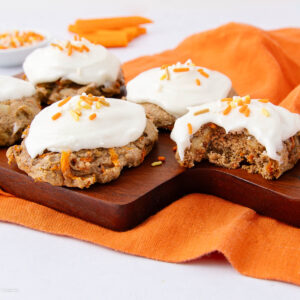 Five carrot cake cookies with white frosting and orange sprinkles sit on a wooden board. One cookie has a bite taken out. An orange cloth and carrot sticks are in the background.
