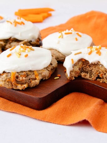 Five carrot cake cookies with white frosting and orange sprinkles sit on a wooden board. One cookie has a bite taken out. An orange cloth and carrot sticks are in the background.