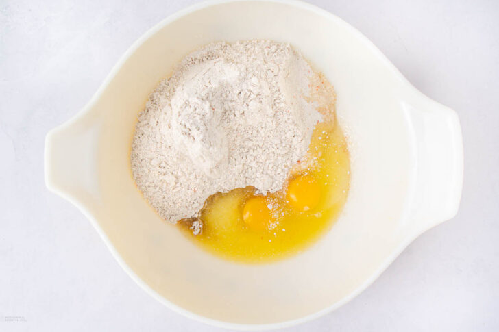 A white mixing bowl containing flour, eggs, and melted butter on a white surface, ready to be mixed for baking.